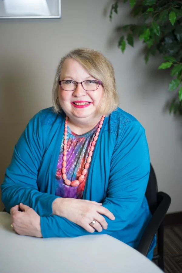 A woman with blonde hair and glasses sitting in front of a wall.