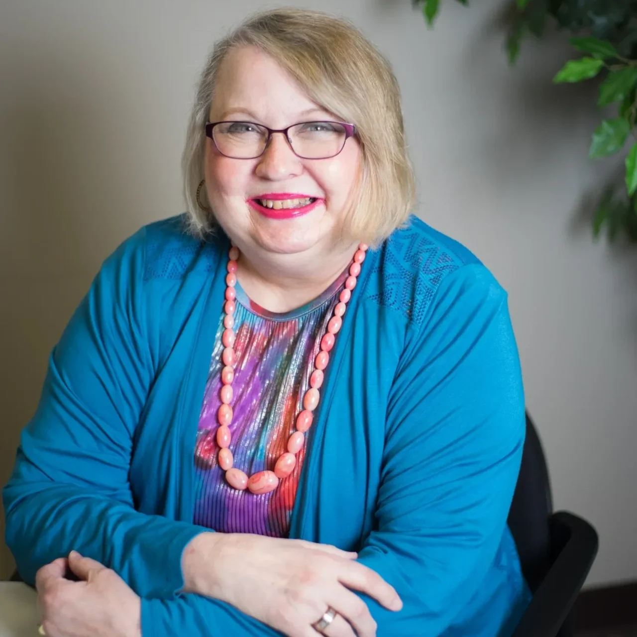 A woman with blonde hair and glasses sitting in front of a wall.